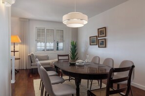 A dining room with a round table, six chairs, a chandelier, a sofa, a window with blinds, and framed pictures on the wall. - Moradia Padre Laurindo – Funchal Bay Views (Funchal)