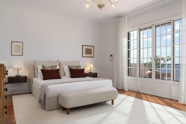 Een slaapkamer met een groot bed, twee nachtkastjes, een kroonluchter en uitzicht door een groot raam.