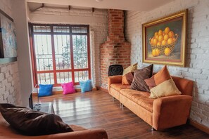A living room with a brick fireplace, a sofa with cushions, a television, and a painting of fruit.