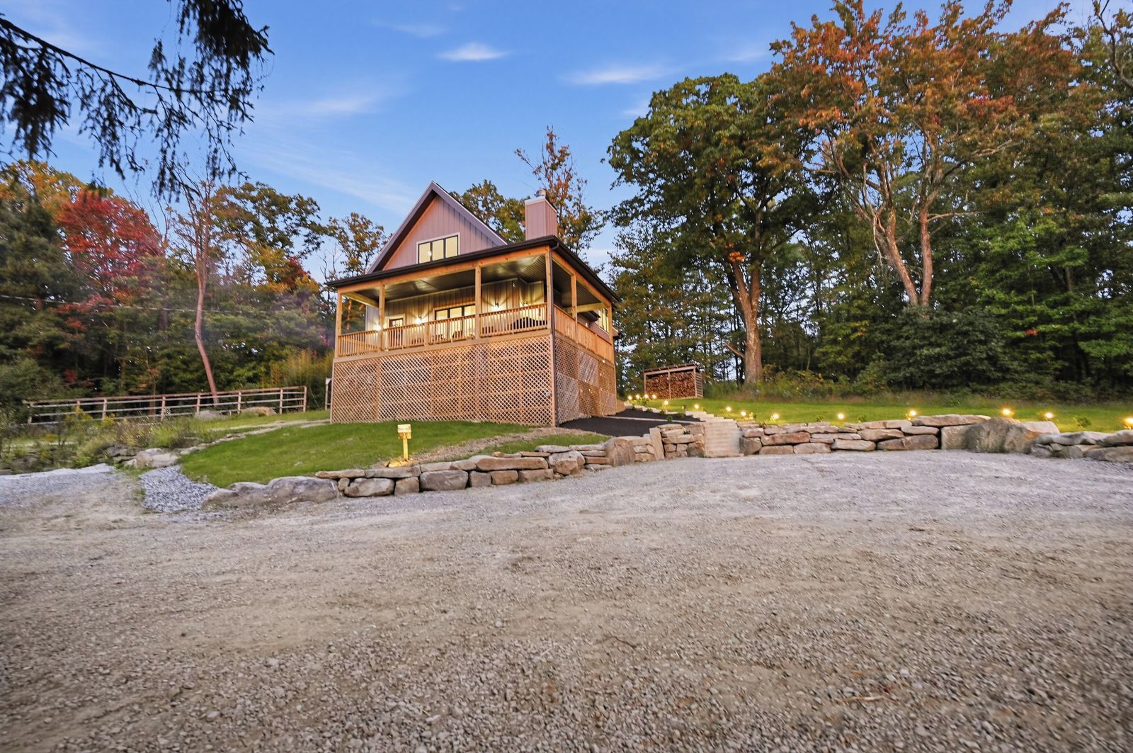 Un chalet rustique doté d’une terrasse en bois et de murs en pierre, entouré d’une allée en gravier et d’un sentier en pierre menant à l’entrée.