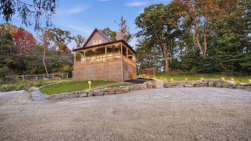 Un chalet rustique doté d’une terrasse en bois et de murs en pierre, entouré d’une allée en gravier et d’un sentier en pierre menant à l’entrée.