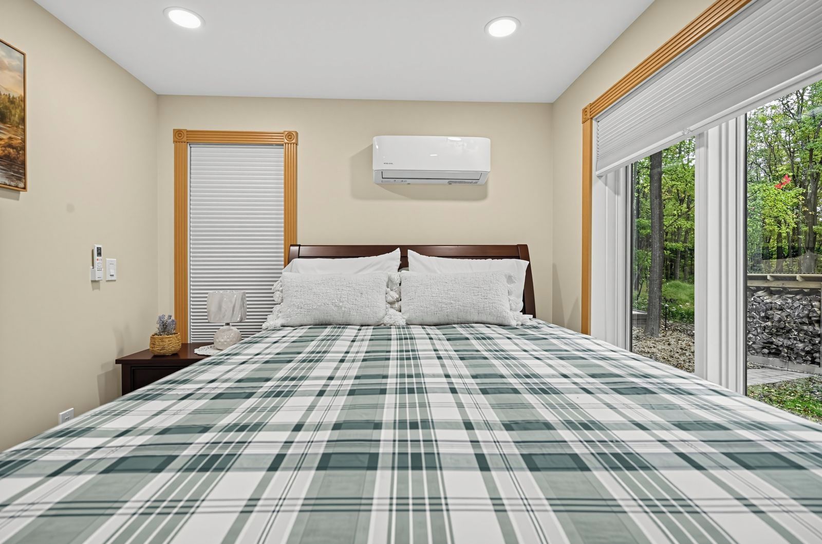 A bedroom with a large bed, a window with blinds, an air conditioning unit, and a view of greenery outside.