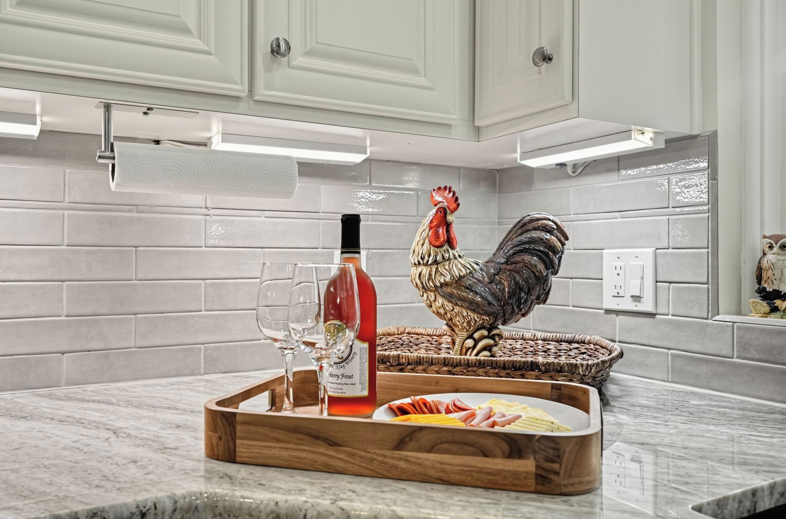 Una encimera de cocina con una bandeja de madera que contiene una botella, vasos y un gallo decorativo.