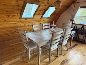 Een houten eettafel met zes stoelen, een kroonluchter en twee dakramen in een kamer met houten wanden en vloeren.