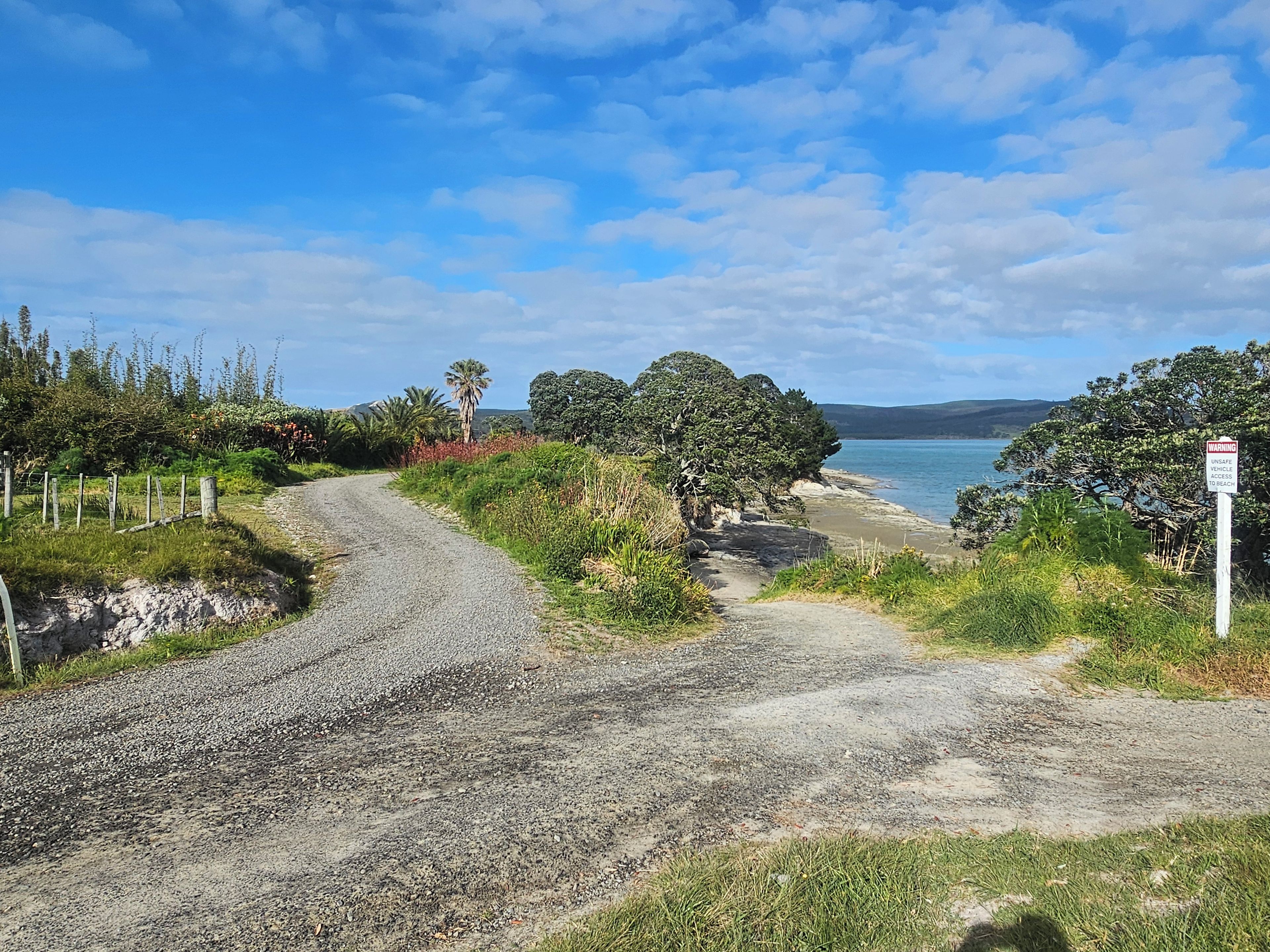 A gravel path leading to a beach with a warning sign, surrounded by greenery and trees.