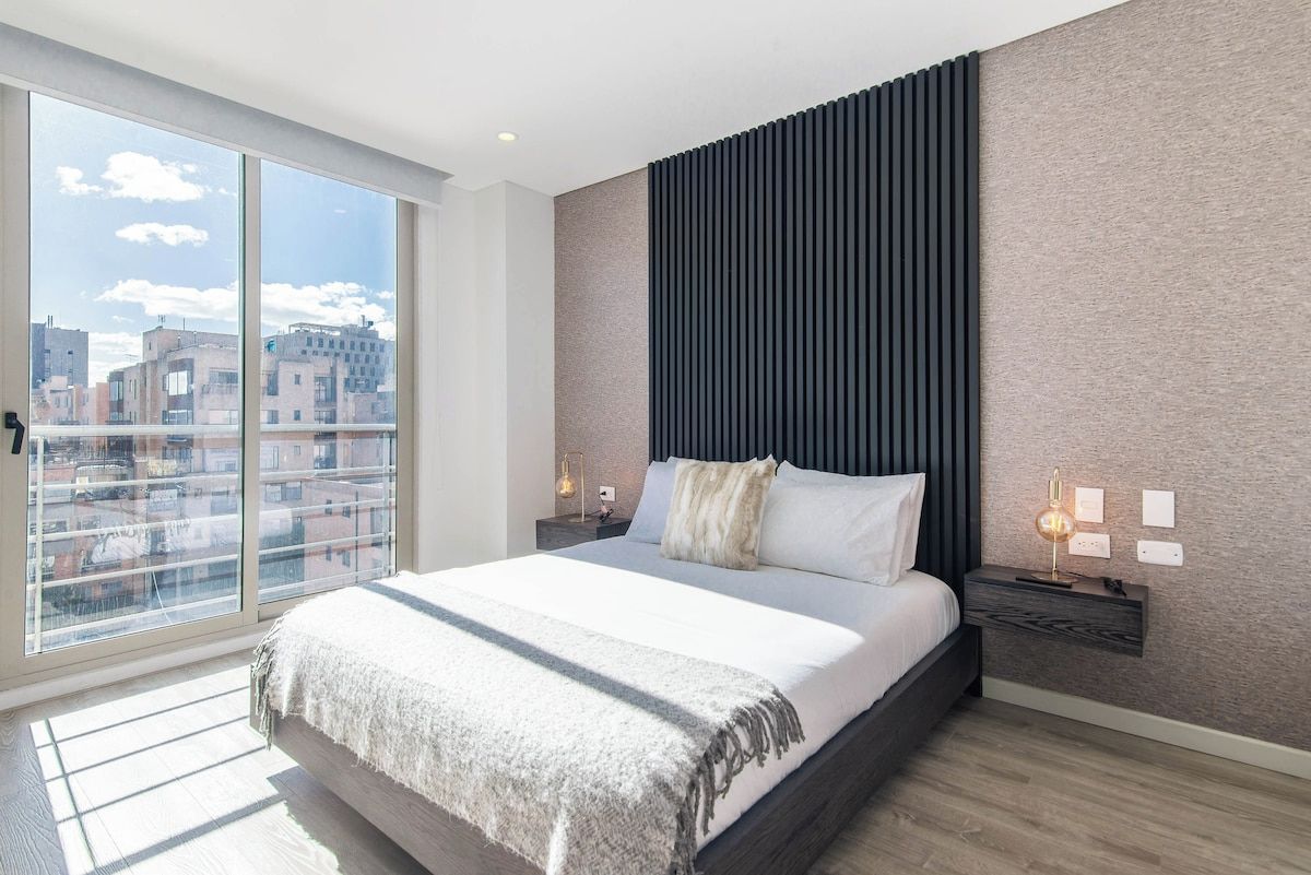 A modern bedroom with a large bed, a city view through floor-to-ceiling windows, a textured wall, and a nightstand with a lamp.