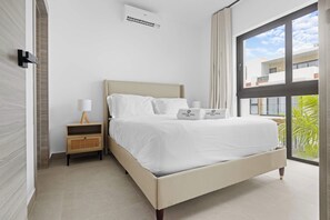 Quarto moderno com cama, mesa de cabeceira e uma grande janela com vista para edifícios e áreas verdes.