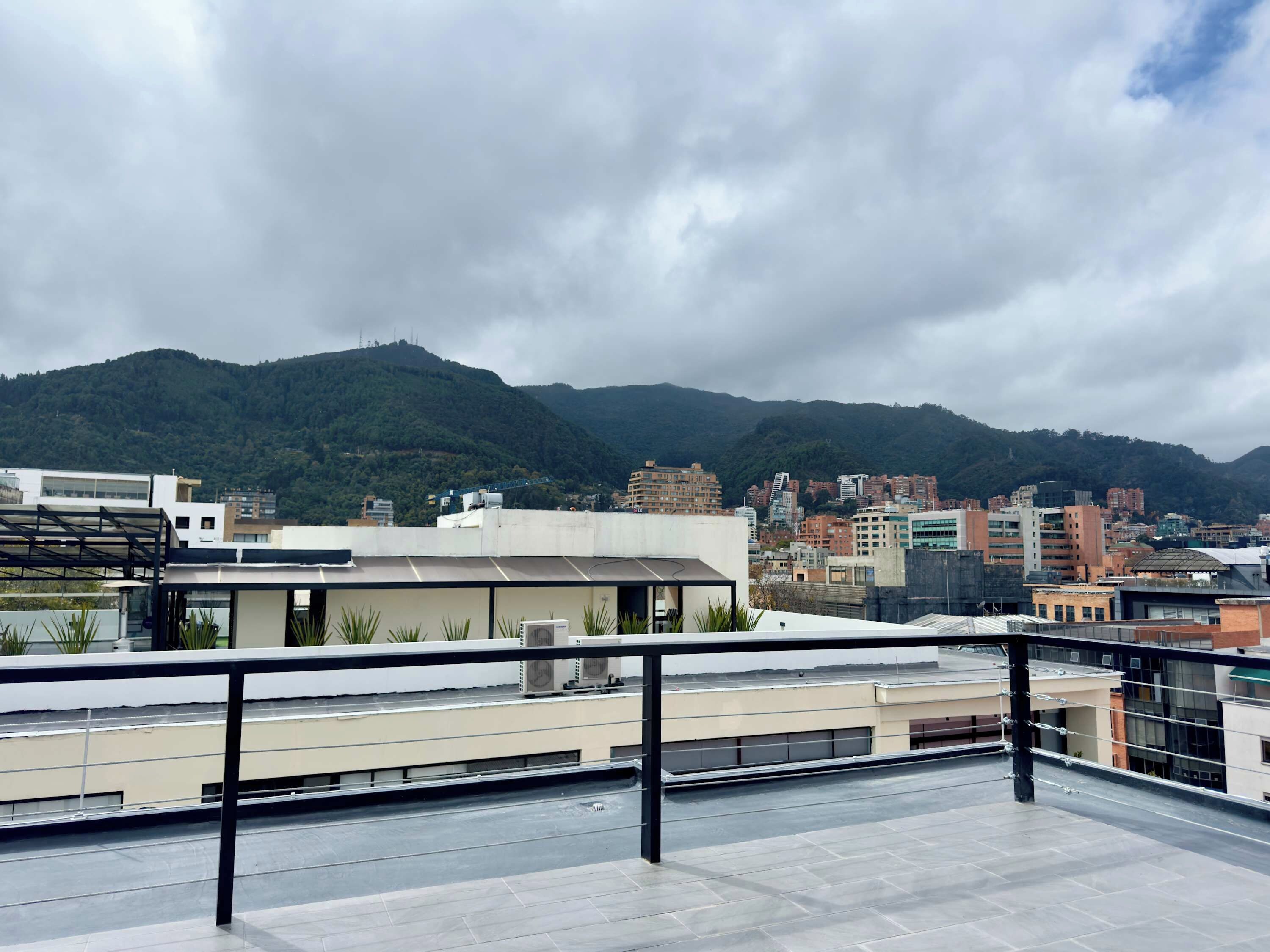 Una terraza en la azotea con barandilla de metal, con vistas a un paisaje urbano que incluye edificios y una montaña al fondo.