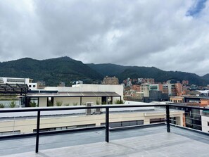 Une terrasse sur le toit, dotée d’une rambarde en métal, qui offre une vue sur un paysage urbain avec des bâtiments et une montagne en arrière-plan.