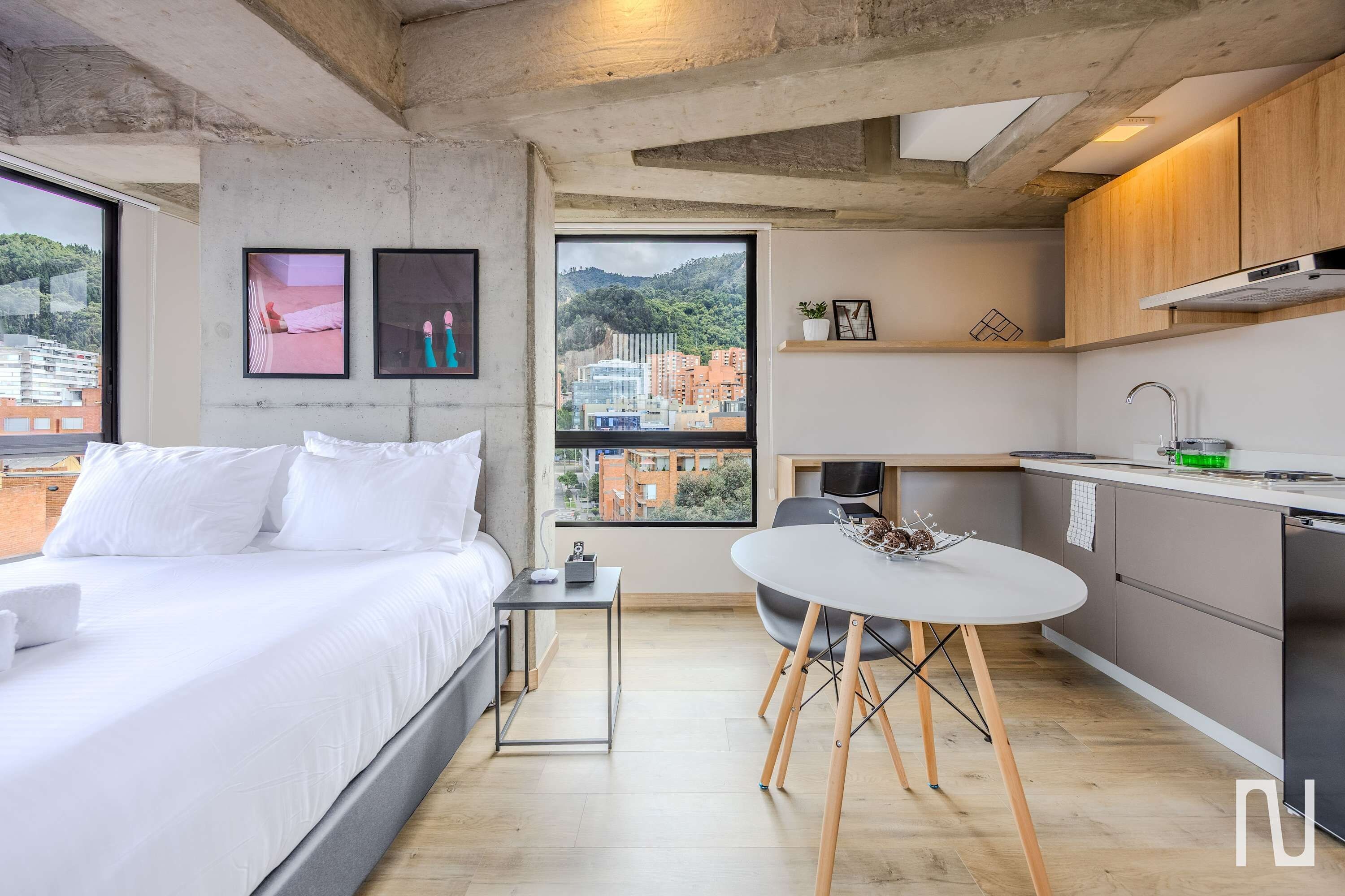 Un hotel moderno con cama, mesita de noche y vistas a un paisaje urbano a través de amplias ventanas.