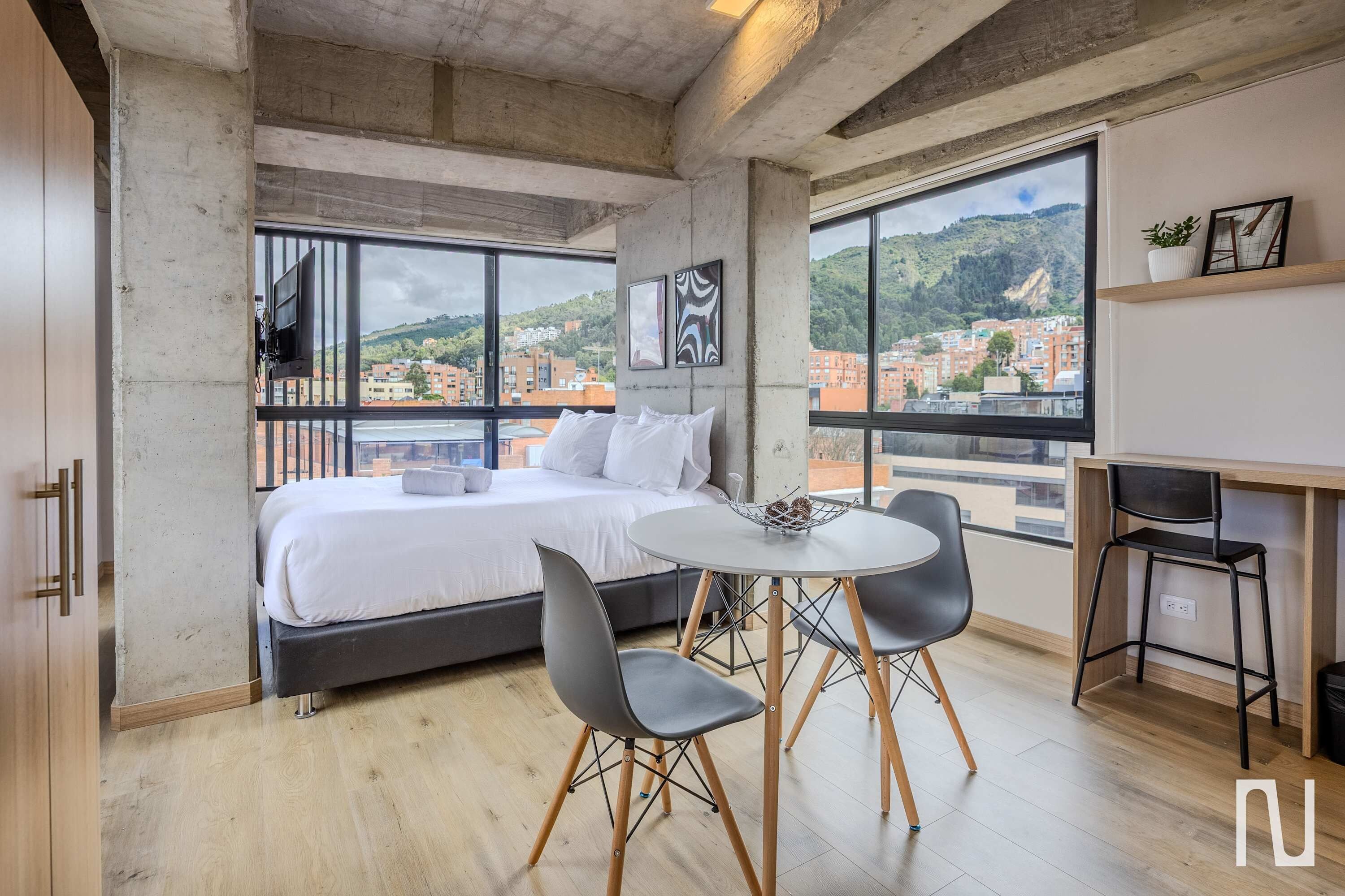 Un dormitorio moderno con una cama grande, una mesa de comedor y sillas, un escritorio pequeño y ventanas amplias con vista a la ciudad.