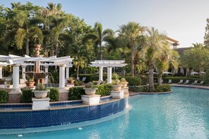 Un área de piscina con una pérgola blanca, palmeras, sillas de descanso y un borde de piscina con mosaicos.