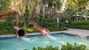 Una piscina con dos toboganes acuáticos, rodeada de exuberante vegetación y un paisaje rocoso.