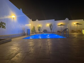 Uma área de piscina externa com piso de azulejos, cercada por paredes com luminárias e cactos.