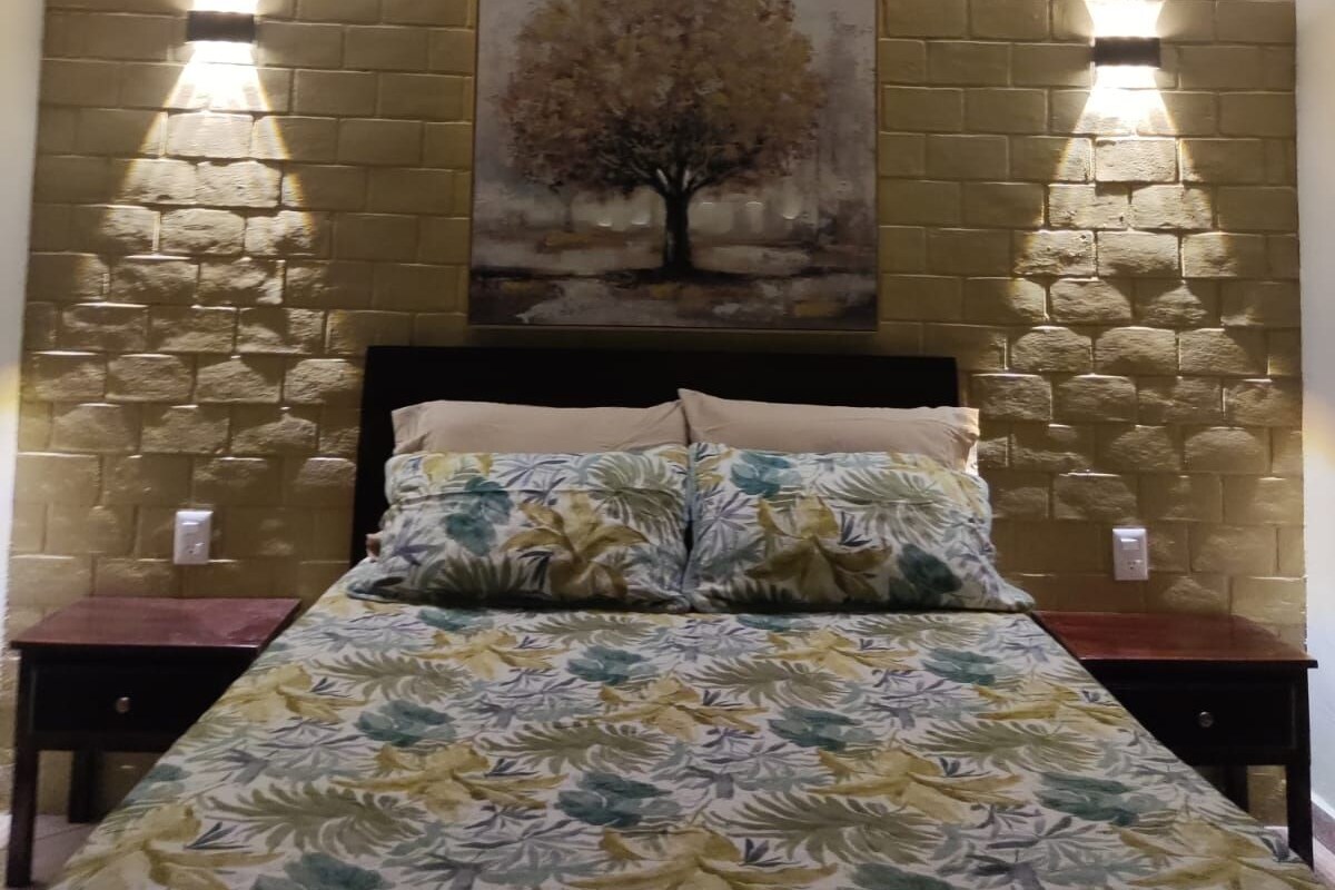 Une chambre à coucher comprenant un lit, un ventilateur de plafond, un tableau représentant un arbre accroché au mur, et deux lampes fixées au mur.