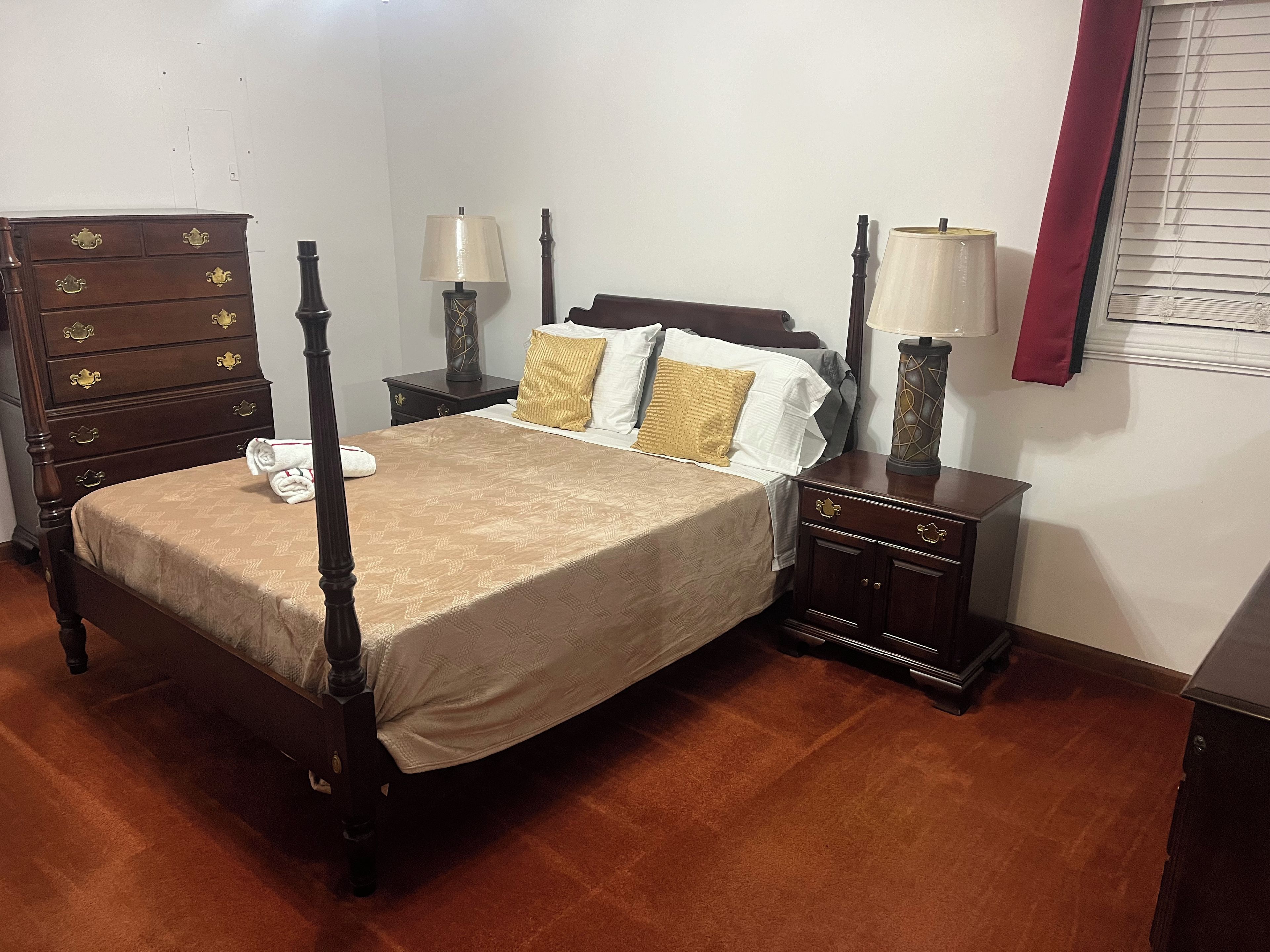 Un lit à baldaquin avec une couvre-lit beige, deux tables de chevet avec des lampes, une commode en bois et une fenêtre avec des stores.