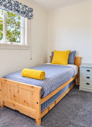 A single bed with a wooden frame, a blue striped bedspread, and a yellow pillow. There is a small bedside table with drawers and a window with a white frame and a blue floral curtain.