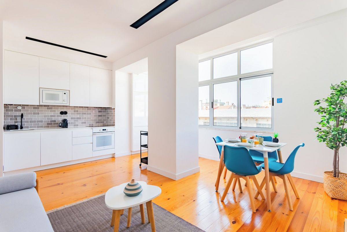 A modern kitchen with white cabinets, a microwave, and a brick backsplash. There's a dining area with blue chairs and a table, and a large window offering a city view.