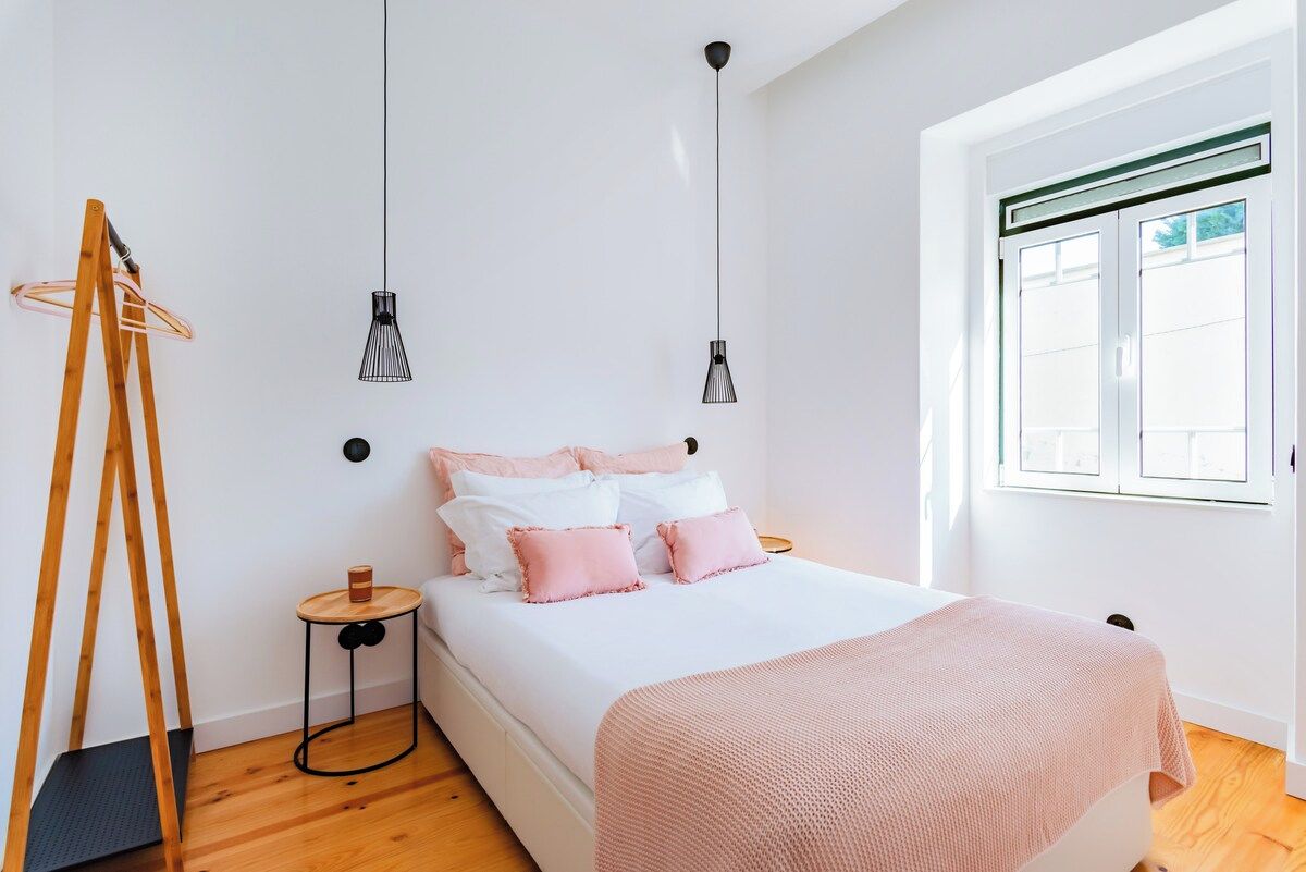 A bedroom with a bed, two pendant lights, a wooden ladder, a small round table, and a window with blinds.