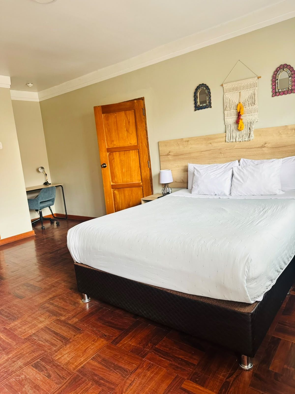 A hotel room with a large bed, wooden flooring, a desk with a chair, and a door.