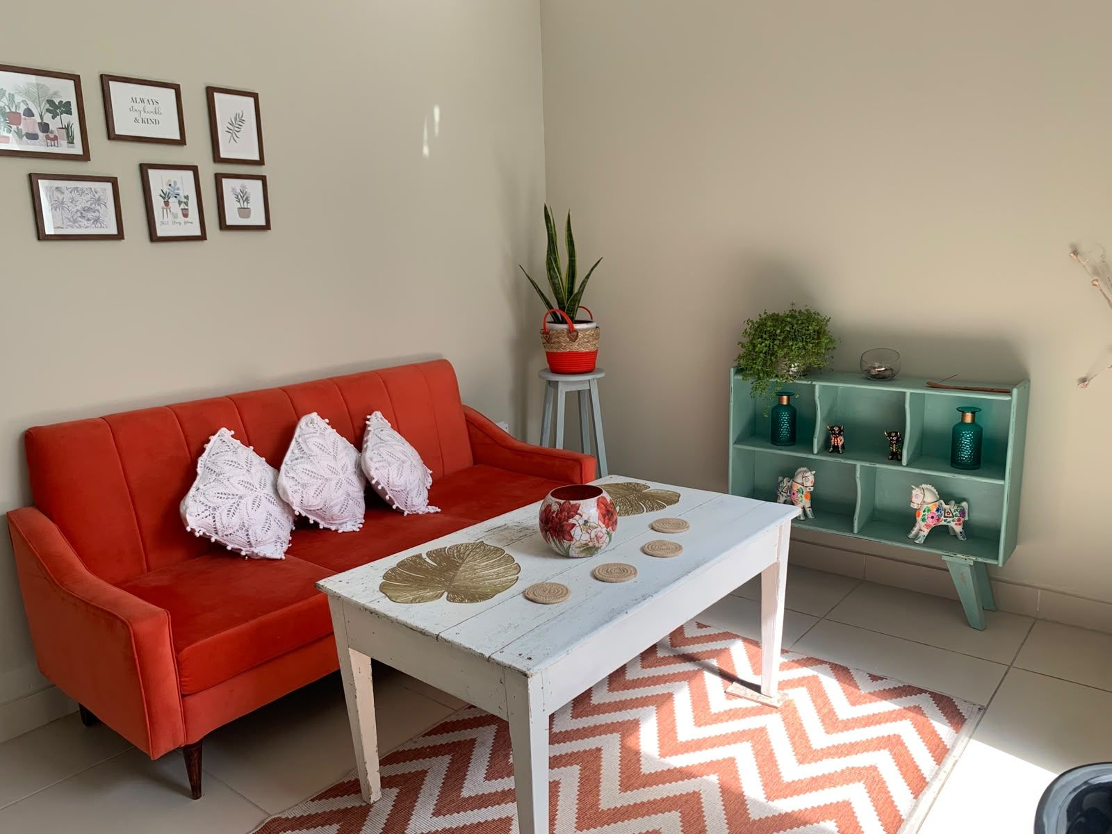 A living room with a red sofa, white cushions, a white coffee table with decorative items, a patterned rug, and a teal shelving unit with small figurines.