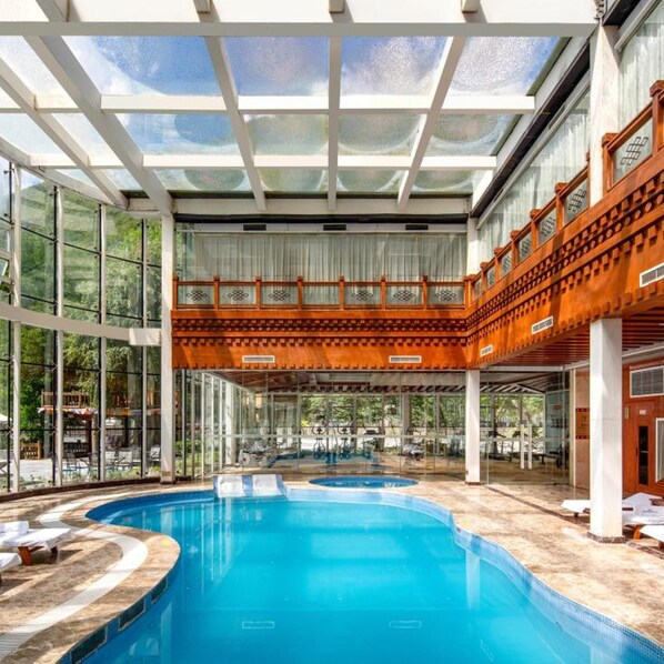 Piscina com teto de vidro, rodeada por espreguiçadeiras e um deck de madeira.