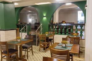 Een restaurantinterieur met houten tafels en stoelen, groene bogen en een trap naar een ander gedeelte.