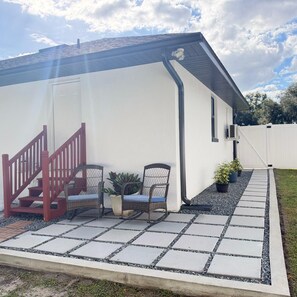 Et hus med hvide facader, en rød trappe, to fletstole med hynder, og en belagt terrasse.