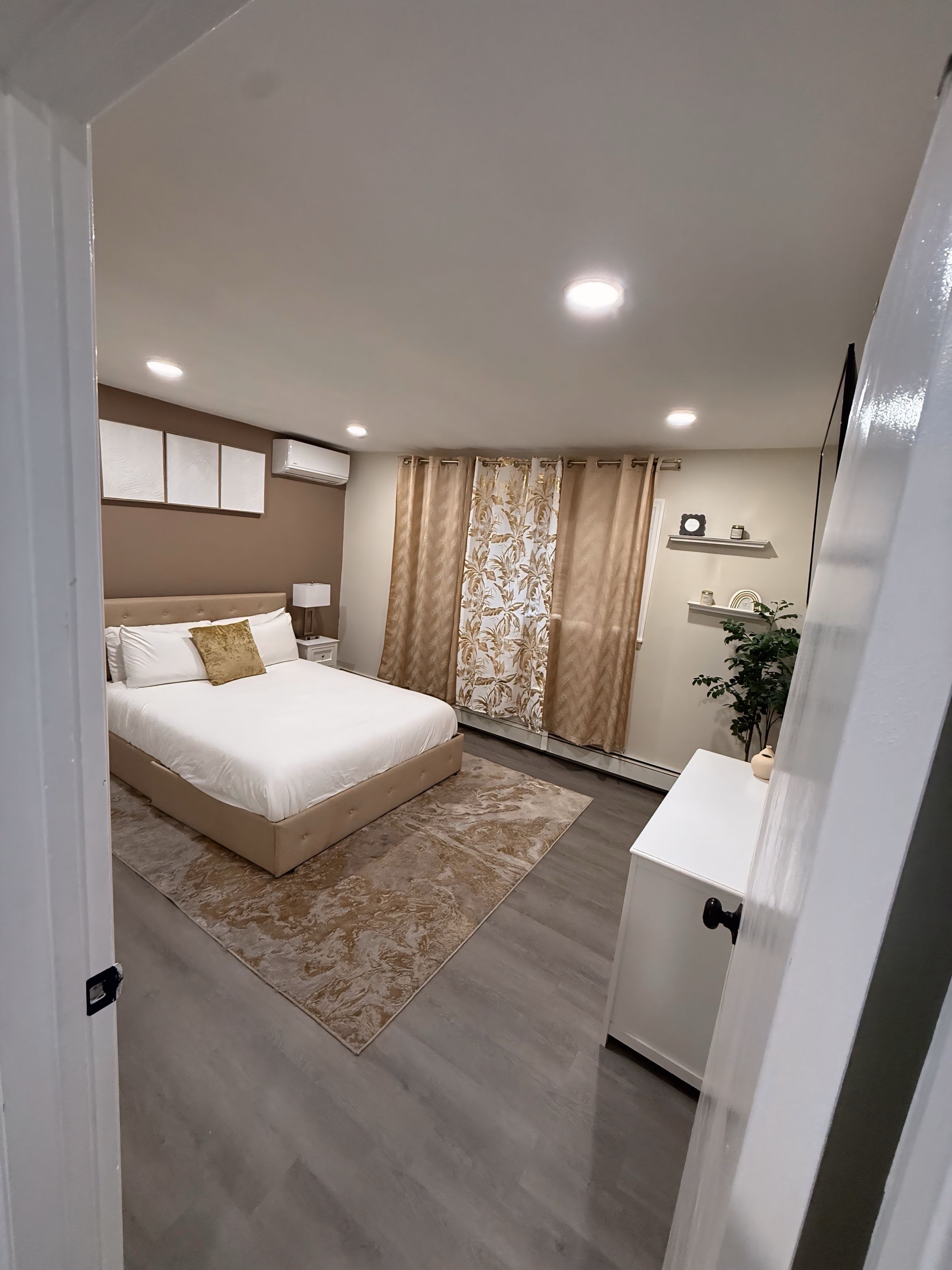 Une chambre à coucher comprenant un lit, des tables de chevet, une commode, un meuble à tiroirs, un miroir, une fenêtre avec des rideaux et une étagère murale agrémentée d’objets décoratifs.