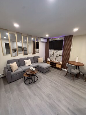 Un salon moderne avec un canapé d’angle gris, une table basse ronde et un coin repas comprenant une petite table et des chaises.