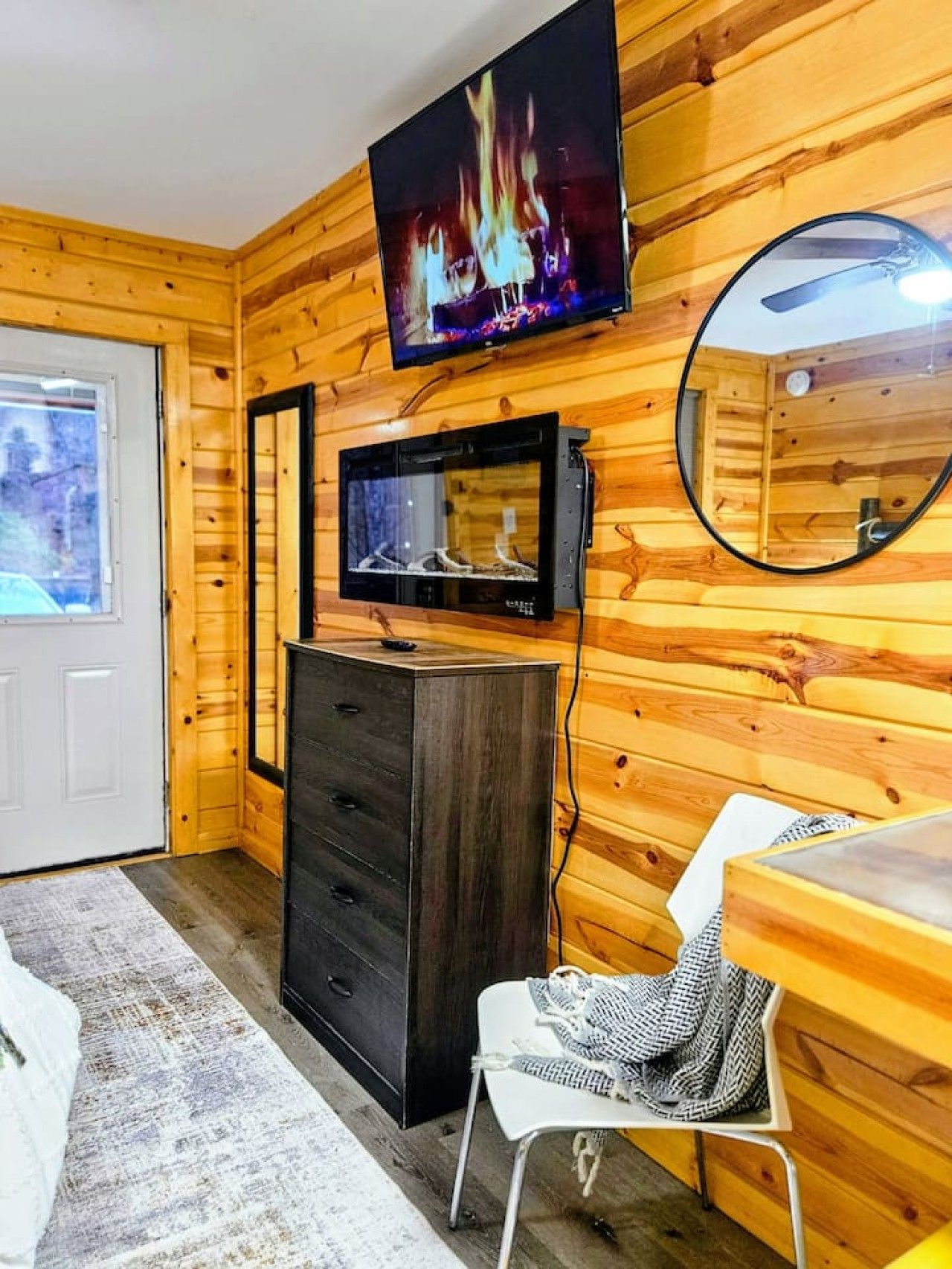 A wooden interior with a television mounted above a dark wooden dresser, a white chair with a patterned cushion, and a round mirror on the wall.