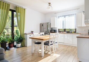 Une cuisine moderne avec des meubles blancs, une table à manger en bois et une vue sur l’extérieur grâce à de grandes fenêtres.