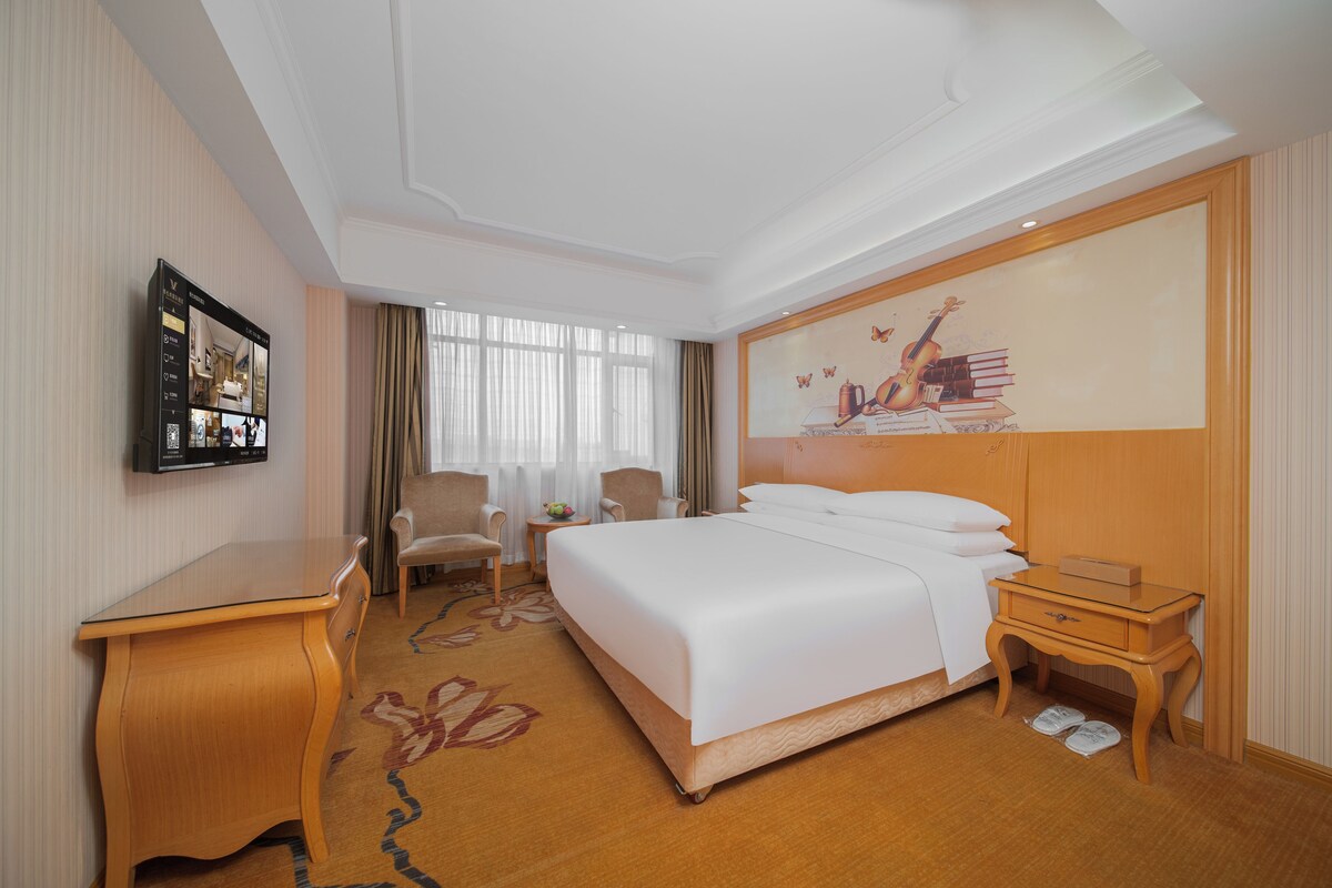 A hotel room with a large bed, a desk, a chair, a small table, and a wall-mounted TV.