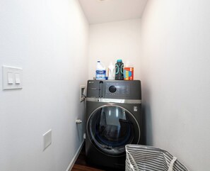 A compact laundry room with a front-loading washing machine, a drying rack, and a small storage area with cleaning supplies. - New Spacious Luxe Home | Views & Game Room! (Tacoma)