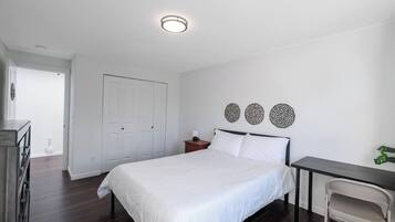 A bedroom with a bed, bedside table, and a desk with a chair. There is a door and a plant in the room.