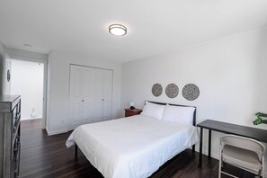 Een slaapkamer met een bed, nachtkastje en bureau met stoel. Er is een deur en een plant in de kamer.