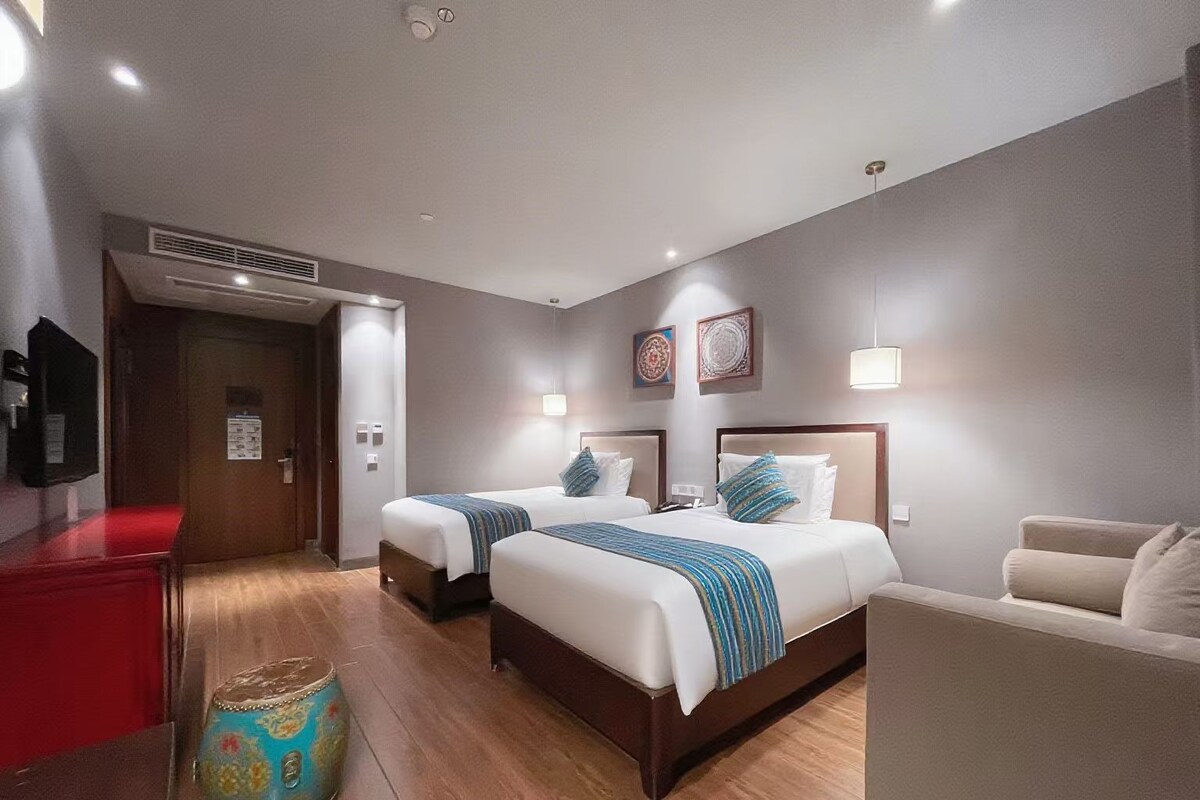 Quarto de hotel com duas camas, um minibar vermelho, piso de madeira e um banquinho decorativo.