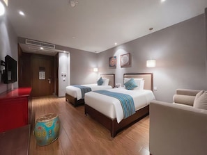 Quarto de hotel com duas camas, um minibar vermelho, piso de madeira e um banquinho decorativo.