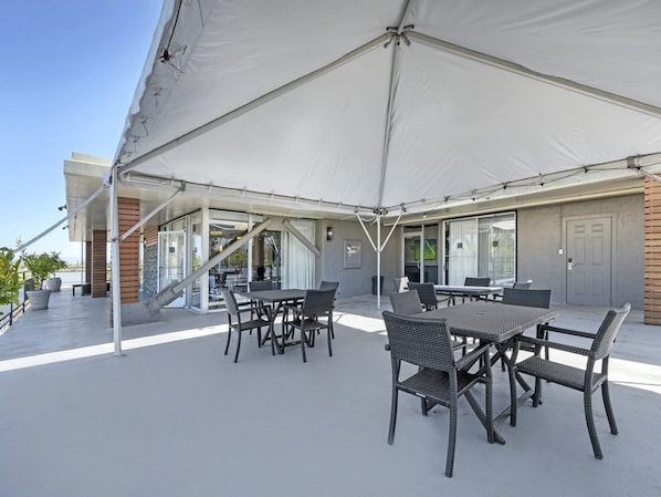 Een terras buiten met een grote witte markies, rieten meubels en een modern gebouw met glazen deuren en ramen.