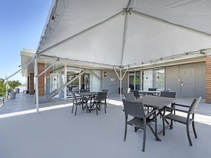Een terras buiten met een grote witte markies, rieten meubels en een modern gebouw met glazen deuren en ramen.