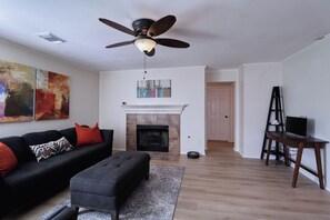 Ein Wohnzimmer mit Kamin, einem schwarzen Sofa mit roten Kissen, einem grauen Hocker, einem Deckenventilator und einem Gemälde an der Wand.