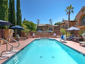 Una piscina con borde de azulejos, rodeada de sillones y sombrillas, con un edificio al fondo.