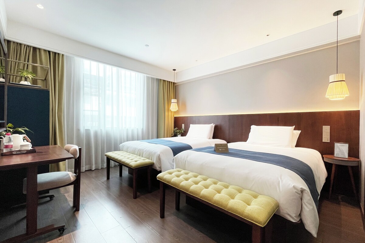 Quarto de hotel com duas camas, uma escrivaninha, um banco e uma mesa de cabeceira.