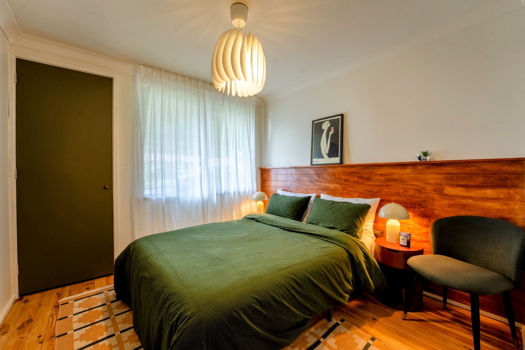 Un dormitorio con cama, una silla, mesita de noche, lámpara y ventana con cortinas.