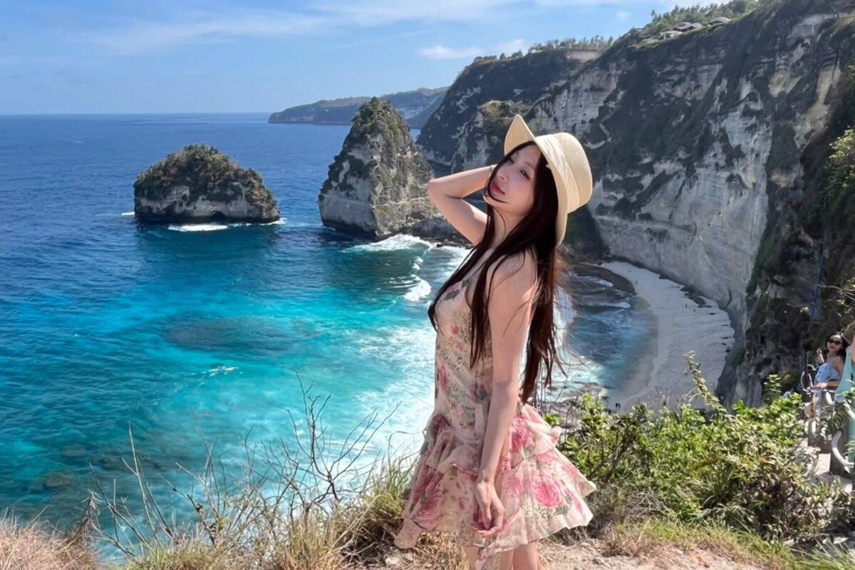 Paisagem costeira com águas azul-claras, falésias rochosas e uma pessoa com vestido e chapéu florais.