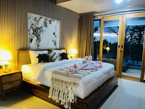 Quarto de hotel com uma cama grande, mesa de cabeceira com abajur, pintura na parede e porta de vidro deslizante para área externa.