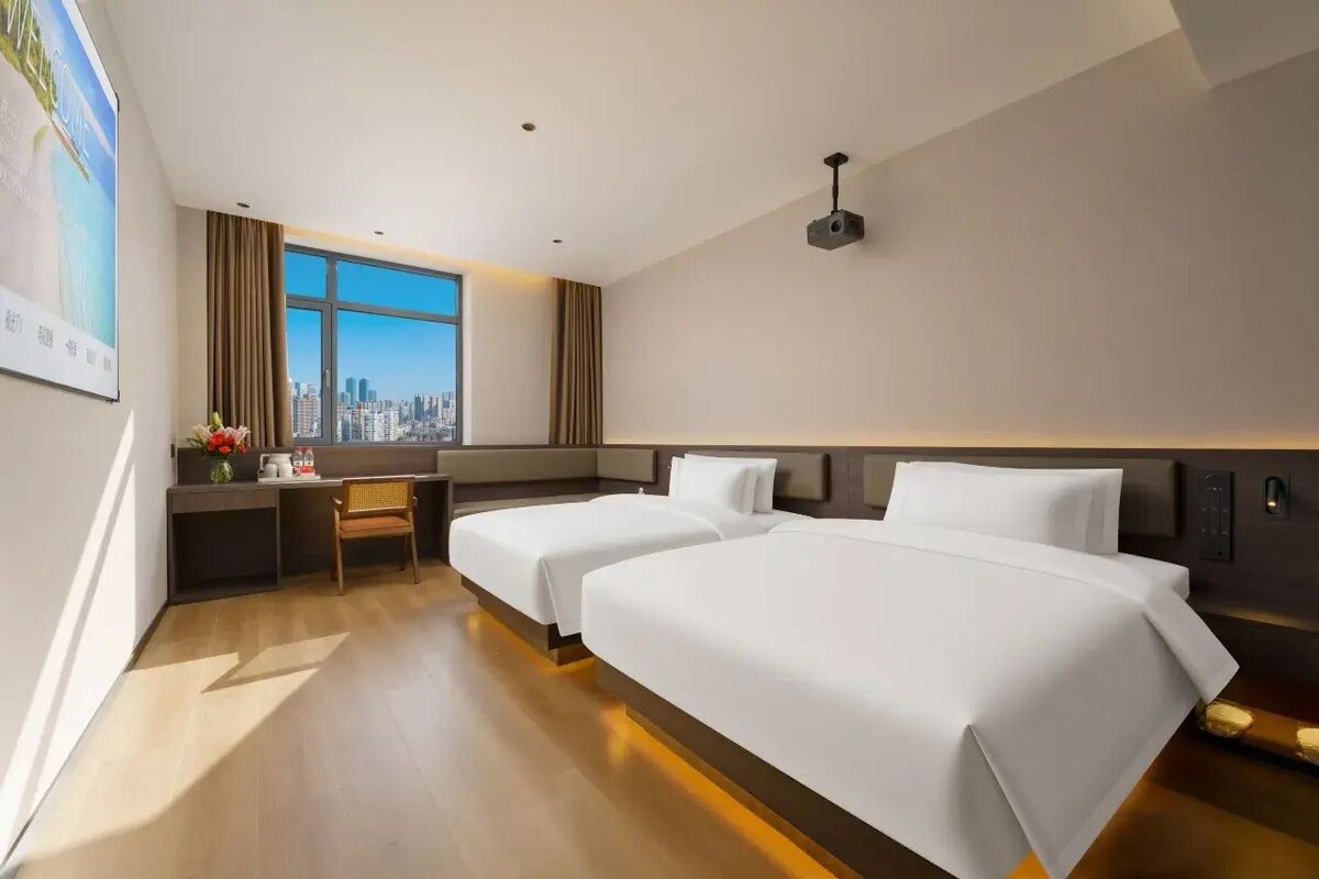 Une chambre d’hôtel avec deux lits, un bureau, une chaise, une fenêtre donnant sur la ville et un projecteur fixé au mur.