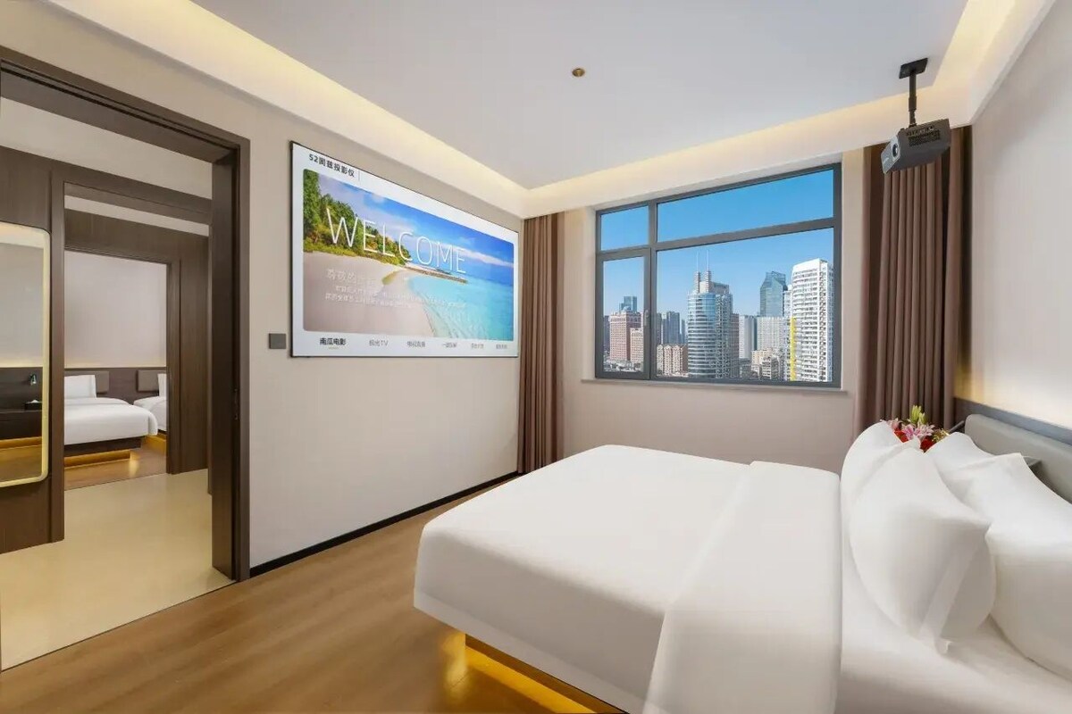 Une chambre d’hôtel avec un lit, un écran de télévision affichant un message de bienvenue, une grande fenêtre donnant sur la ville et une porte menant à une autre pièce.