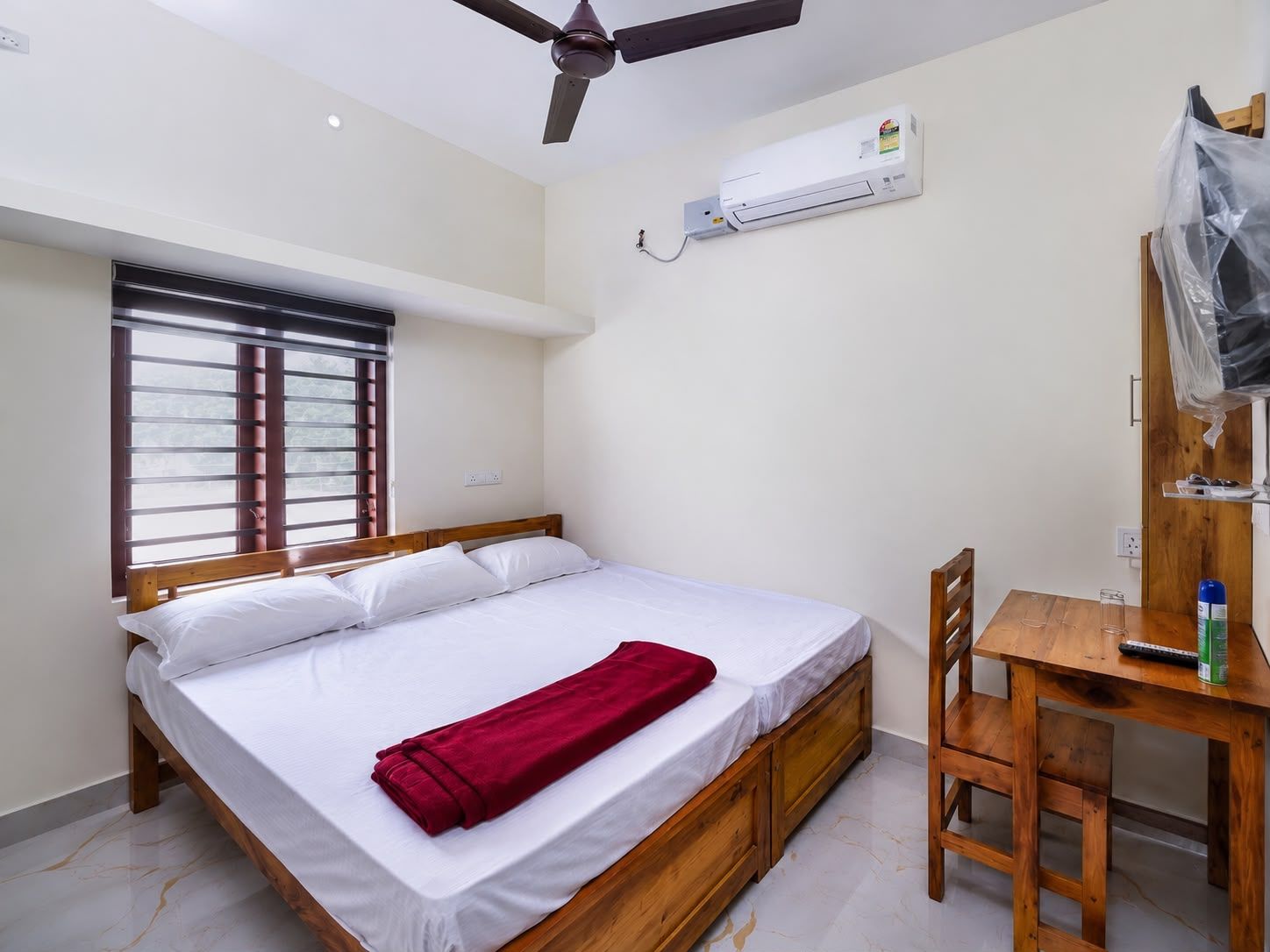Une chambre avec un lit, un ventilateur de plafond, un appareil de climatisation, une fenêtre avec des stores et une table en bois avec une chaise.