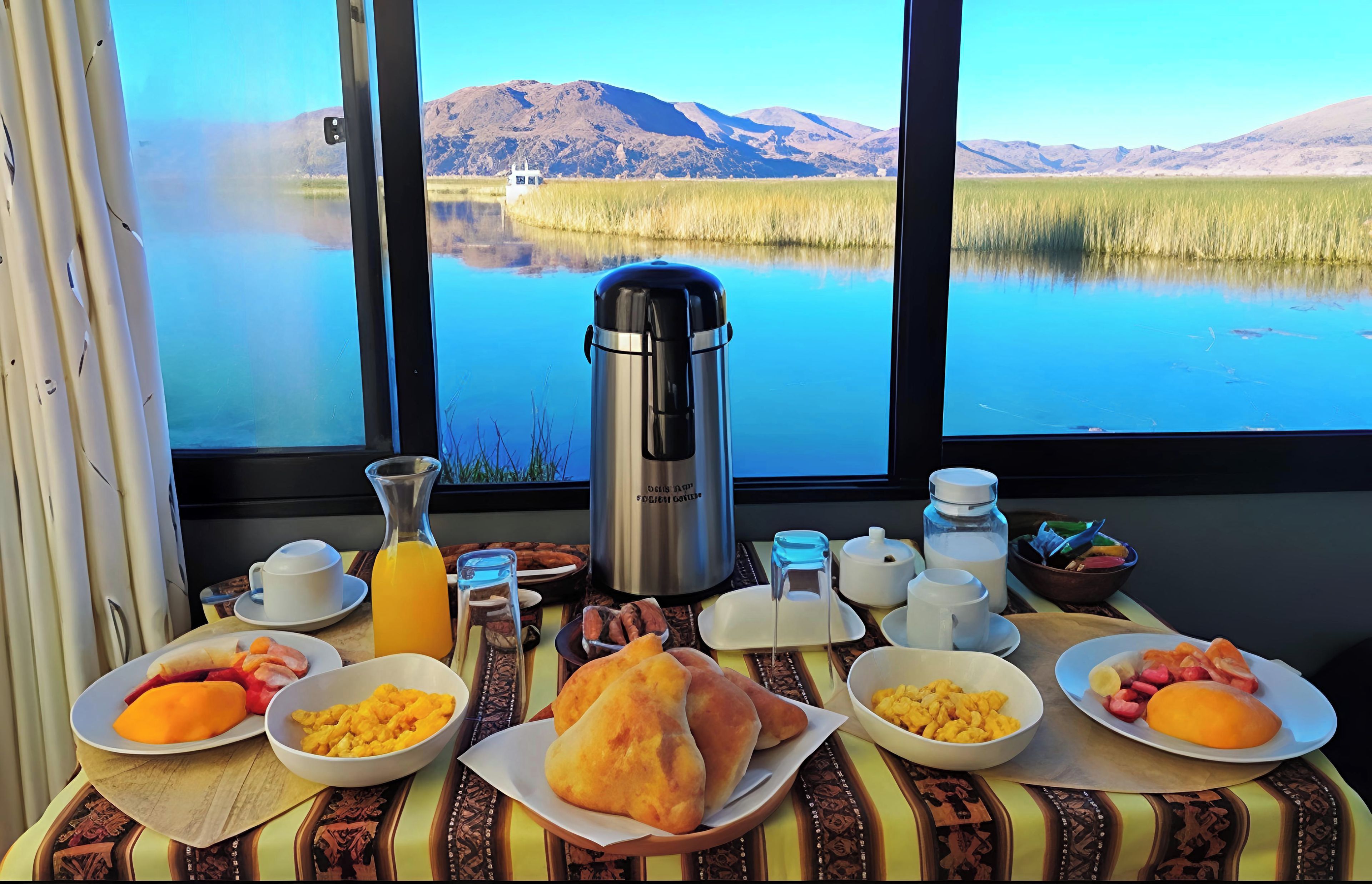 A breakfast table with various food items and a coffee maker, set against a scenic view of a lake and mountains.